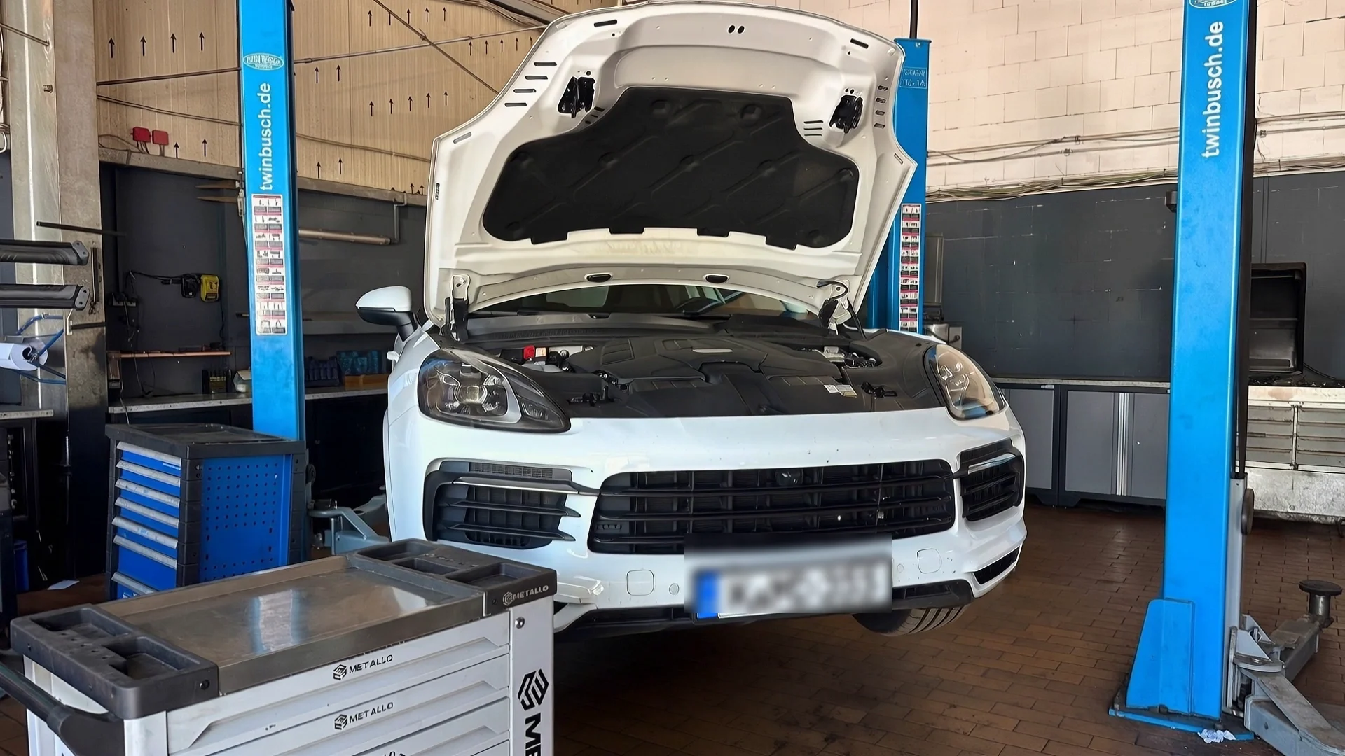 Derselbe Wagen mit geöffneter Motorhaube, von der Hebebühne angehoben — die Räder hängen frei über dem Hallenboden.