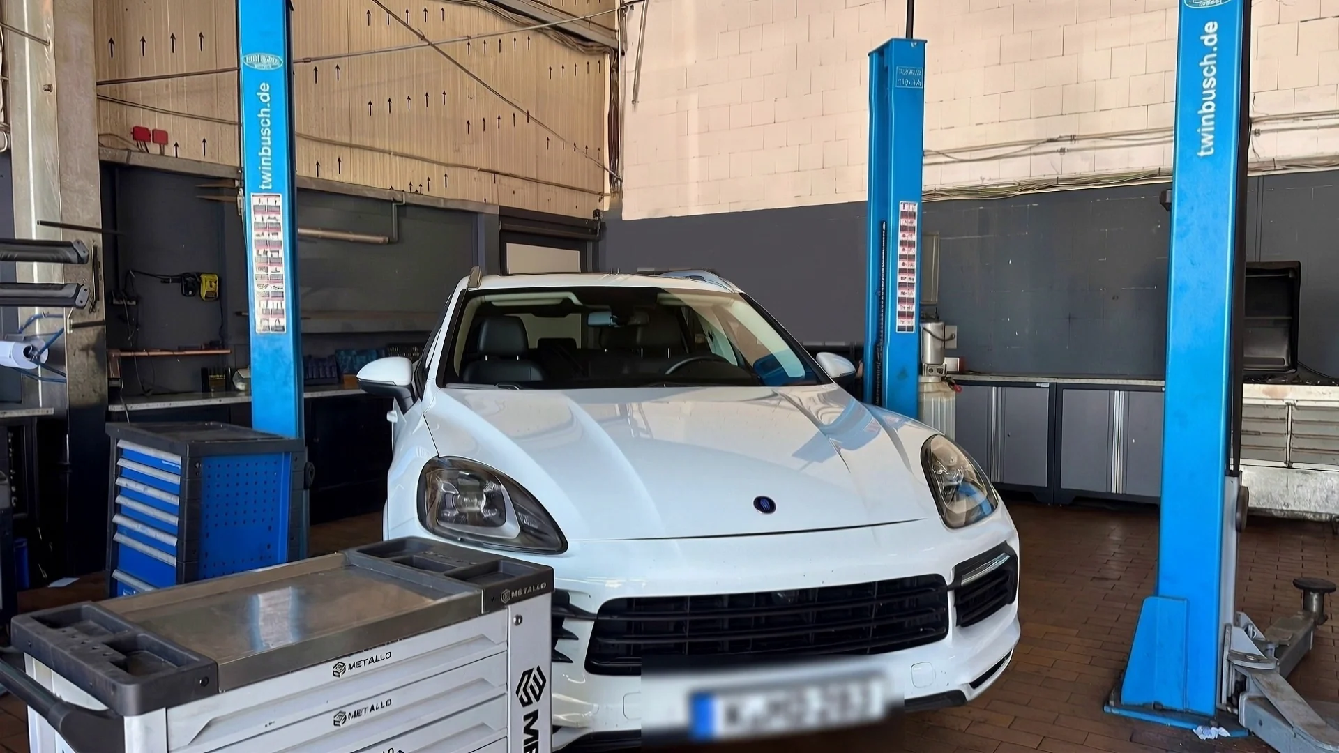 Ein weißer Porsche Cayenne steht mit geschlossener Motorhaube zwischen den Säulen der Hebebühne in der Halle von Boostwerk Köln.